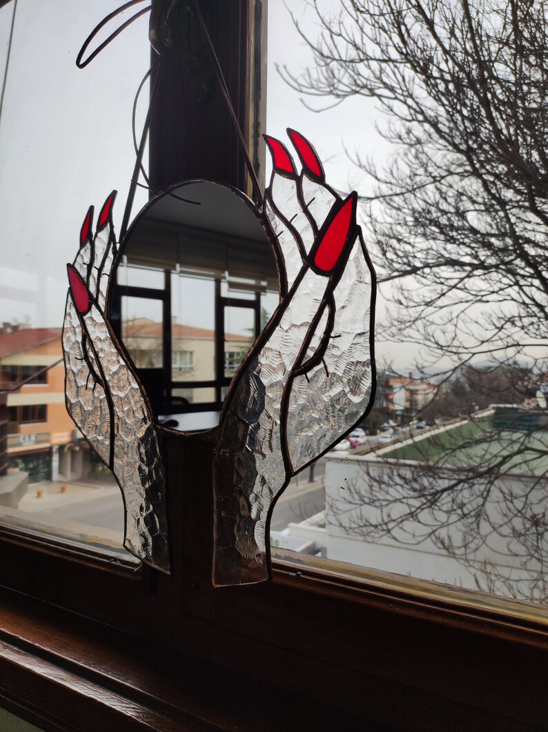 May include: Stained glass mirror in the shape of hands with red fingernails. The hands are holding a round mirror. The glass has a textured appearance. The mirror is hanging in front of a window.