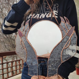 May include: A round mirror framed by stained glass hands with copper-colored edging. The hands have purple accents on the nails and are holding the mirror. A brown leather cord is attached for hanging.