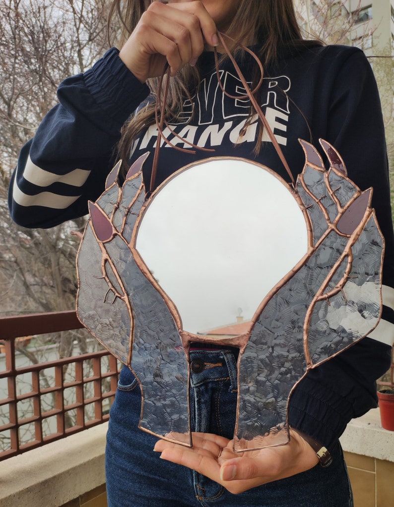 May include: A round mirror framed by stained glass hands with copper-colored edging. The hands have purple accents on the nails and are holding the mirror. A brown leather cord is attached for hanging.
