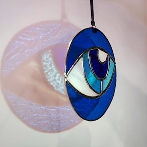 Puede incluir: Un adorno de ojo de vidrio azul y blanco que cuelga de una cuerda negra. El ojo está rodeado por un círculo azul.