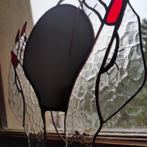 May include: Stained glass art piece depicting hands with red fingernails holding a dark sphere. The glass has a textured, patterned appearance. The piece is framed with dark brown lines, creating a unique decorative item.