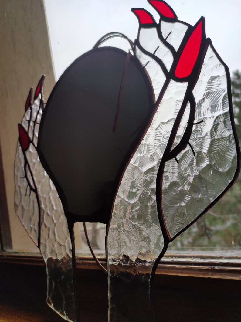 May include: Stained glass art piece depicting hands with red fingernails holding a dark sphere. The glass has a textured, patterned appearance. The piece is framed with dark brown lines, creating a unique decorative item.