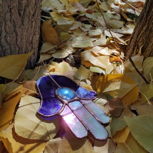 May include: A stained glass Hamsa hand ornament with a blue and iridescent design. The hand-shaped art piece features a central blue gem and is surrounded by a purple and light blue color scheme. The ornament is displayed on a bed of yellow leaves.