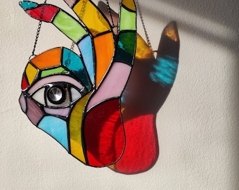 May include: A stained glass suncatcher shaped like a hand with an eye in the palm, featuring multicolored glass segments in red, yellow, blue, green, and pink, held together by black metal solder lines and suspended by a thin black metal chain.