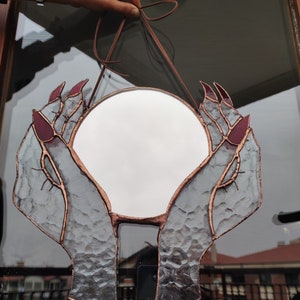 May include: A round stained glass mirror featuring hands with long, burgundy nails cradling the reflective surface. The glass has a textured appearance, with copper-colored lines outlining the hands and mirror. A brown leather cord is attached for hanging.
