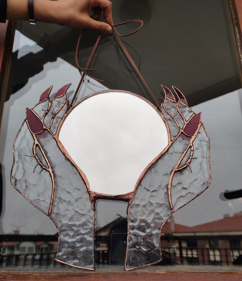 May include: A round stained glass mirror featuring hands with long, burgundy nails cradling the reflective surface. The glass has a textured appearance, with copper-colored lines outlining the hands and mirror. A brown leather cord is attached for hanging.