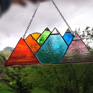 May include: A stained glass mountain range suncatcher with a rainbow of colors, including red, orange, yellow, green, blue, and purple. The suncatcher is hanging from a chain.