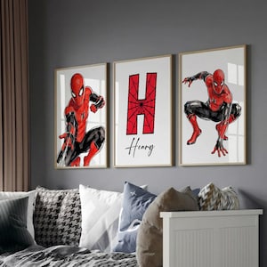 May include: Three framed art prints featuring Spider-Man in action poses. The middle print displays a large red letter "H" with a spiderweb design and the name "Henry" below. The prints are displayed above a white daybed.