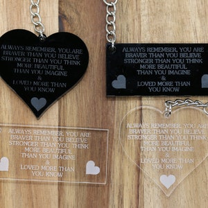 May include: Four keychains with different designs. Each keychain features a heart-shaped or rectangular charm with the text "Always remember, you are braver than you believe stronger than you think more beautiful than you imagine & loved more than you know". Two keychains are black with white text, and two keychains are clear with black text.