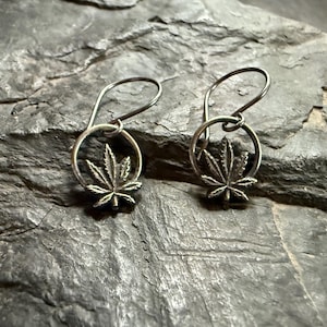 Puede incluir: Un par de pendientes de plata con un diseño de hoja de marihuana. Los pendientes están hechos de un alambre fino y tienen un pequeño círculo en la parte superior.