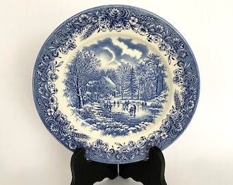 May include: A blue and white decorative plate featuring a winter scene with a house, trees, and people ice skating. The plate has a floral border. The plate is displayed on a black stand.
