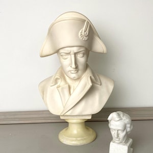 Op de afbeelding: Een cr&egrave;mekleurige buste van een historische figuur met een steek en jas. Een kleinere, witte buste van een andere figuur staat ernaast. De sculpturen staan op een lichtgekleurd oppervlak.