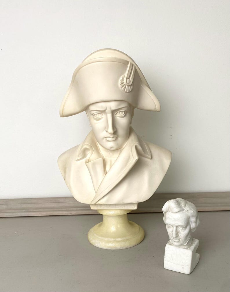 Op de afbeelding: Een cr&egrave;mekleurige buste van een historische figuur met een steek en jas. Een kleinere, witte buste van een andere figuur staat ernaast. De sculpturen staan op een lichtgekleurd oppervlak.