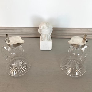 May include: Two clear glass cruet bottles with silver-colored lids and handles. The bottles have a starburst design on the bottom. A small white bust sculpture is between the bottles.