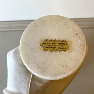 Op de afbeelding: Close-up van een cr&egrave;mekleurig albasten object met een afgeronde bovenkant. Een kleine, rechthoekige sticker met gouden letters is op de bovenkant bevestigd, met de tekst "ALBATRE MOUL&Eacute; retouch&eacute; main D&Eacute;COROP PARIS."
