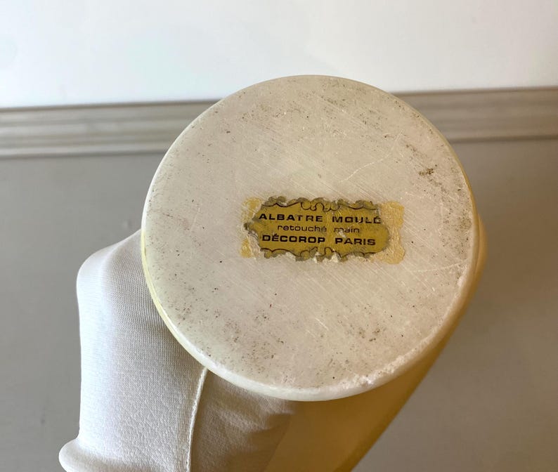 Op de afbeelding: Close-up van een cr&egrave;mekleurig albasten object met een afgeronde bovenkant. Een kleine, rechthoekige sticker met gouden letters is op de bovenkant bevestigd, met de tekst "ALBATRE MOUL&Eacute; retouch&eacute; main D&Eacute;COROP PARIS."