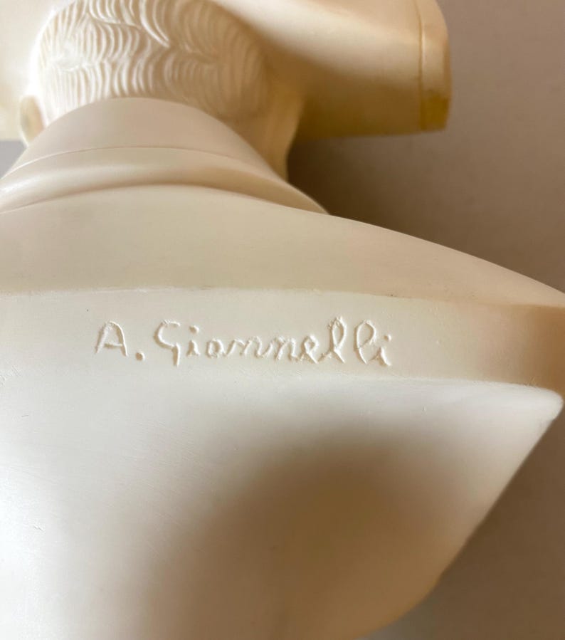 Op de afbeelding: Een cr&egrave;mekleurige borstbeeldsculptuur met gedetailleerde gelaatstrekken en haar. De sculptuur heeft een gladde afwerking en de naam "A. Giannelli" is op de basis gegraveerd. Het borstbeeld is een klassiek kunstwerk.