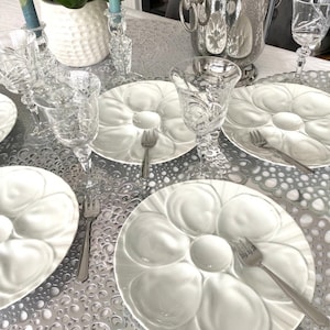 Puede incluir: Una mesa puesta con seis platos de ostras blancos, cada uno con seis hendiduras individuales para servir ostras. Los platos están dispuestos sobre un mantel estampado en plata y blanco. También hay seis copas de vino de vidrio transparente y dos tenedores de plata en la mesa.
