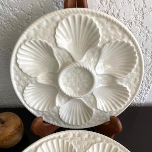 May include: Two white ceramic oyster plates with a raised shell design. The plates are displayed on a wooden stand.
