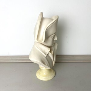 Op de afbeelding: Een cr&egrave;mekleurige buste-sculptuur van een historisch figuur, met een gedetailleerde weergave van een persoon met een hoed en een jas met kraag. De buste is gemonteerd op een ronde, lichtgele basis, tegen een neutrale achtergrond.