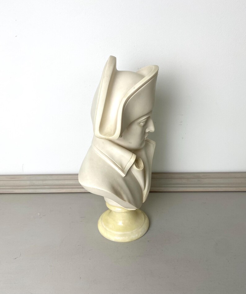 Op de afbeelding: Een cr&egrave;mekleurige buste-sculptuur van een historisch figuur, met een gedetailleerde weergave van een persoon met een hoed en een jas met kraag. De buste is gemonteerd op een ronde, lichtgele basis, tegen een neutrale achtergrond.