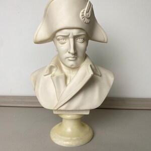 Op de afbeelding: Cr&egrave;mekleurige buste van een man met een steek en een jas in militaire stijl. De sculptuur heeft een gedetailleerd gezicht met een strenge uitdrukking en is op een voetstuk gemonteerd.