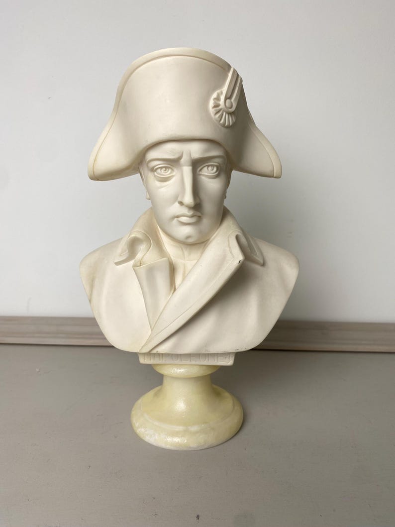 Op de afbeelding: Cr&egrave;mekleurige buste van een man met een steek en een jas in militaire stijl. De sculptuur heeft een gedetailleerd gezicht met een strenge uitdrukking en is op een voetstuk gemonteerd.