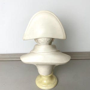 Op de afbeelding: Cr&egrave;mekleurige buste van een historische figuur, mogelijk Napoleon, met een steek. De sculptuur heeft een gedetailleerd gezicht en is gemonteerd op een geel getinte sokkel. De buste is gemaakt van een massief materiaal.