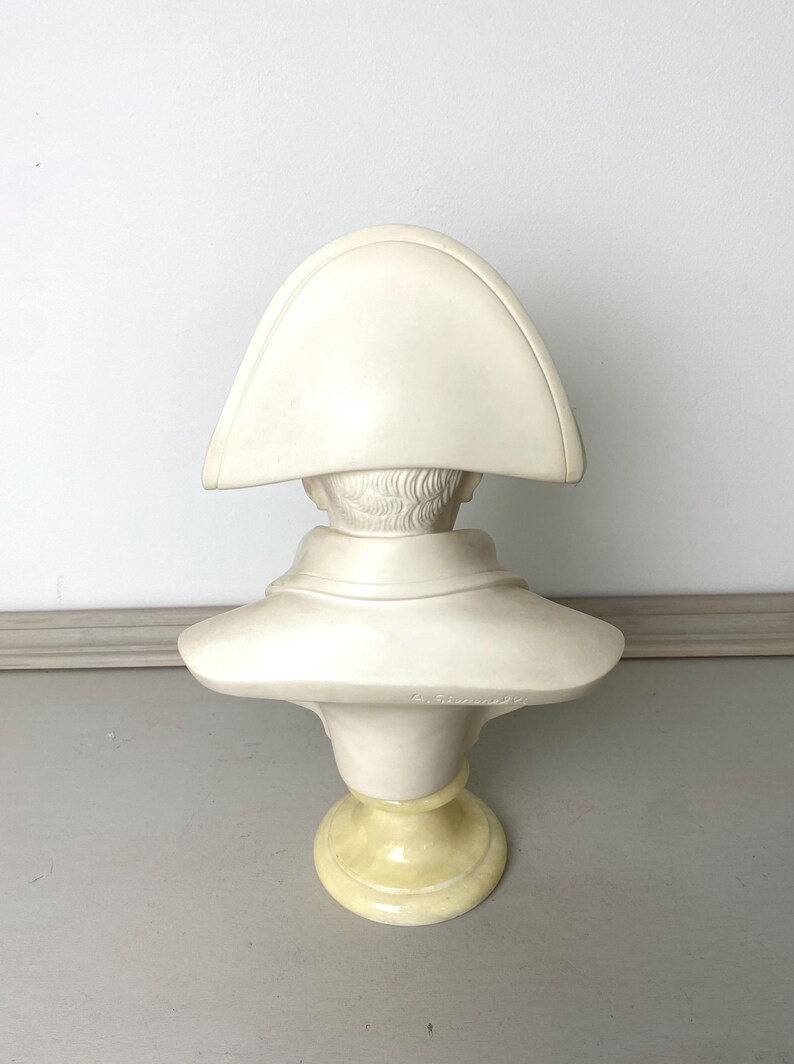 Op de afbeelding: Cr&egrave;mekleurige buste van een historische figuur, mogelijk Napoleon, met een steek. De sculptuur heeft een gedetailleerd gezicht en is gemonteerd op een geel getinte sokkel. De buste is gemaakt van een massief materiaal.