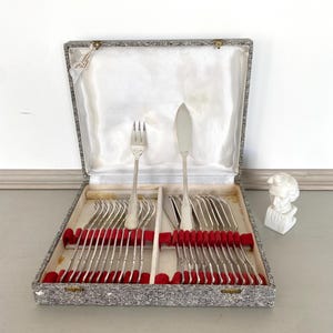 Peut inclure: Un ensemble de couverts argentés vintage dans une boîte grise à motif tweed avec une doublure en velours rouge. L'ensemble comprend des fourchettes, des couteaux et des cuillères. La boîte est ouverte, révélant l'argenterie bien rangée. Un petit buste blanc est à droite.