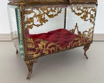 May include: An antique glass and brass display box with a red velvet interior. The box features ornate gold-colored floral detailing and rests on delicate, curved legs. The hinged lid is trimmed with a green border.