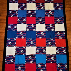 May include: A patchwork quilt featuring a repeating pattern of red, white, blue, and maroon squares. The blue squares display a football helmet logo with the word "BILLS". The quilt is bordered with a black fabric.