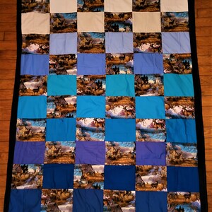 Puede incluir: Una colcha de retazos con un borde negro. La colcha está hecha de cuadrados de diferentes tonos de azul, blanco y turquesa. Cada cuadrado presenta una imagen impresa de un ciervo en un entorno boscoso.