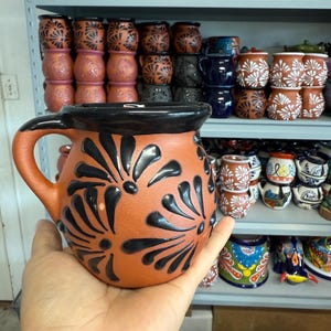 Puede incluir: Una jarra de terracota con un borde negro y dise&ntilde;os florales negros. La jarra tiene un asa y est&aacute; hecha de arcilla.
