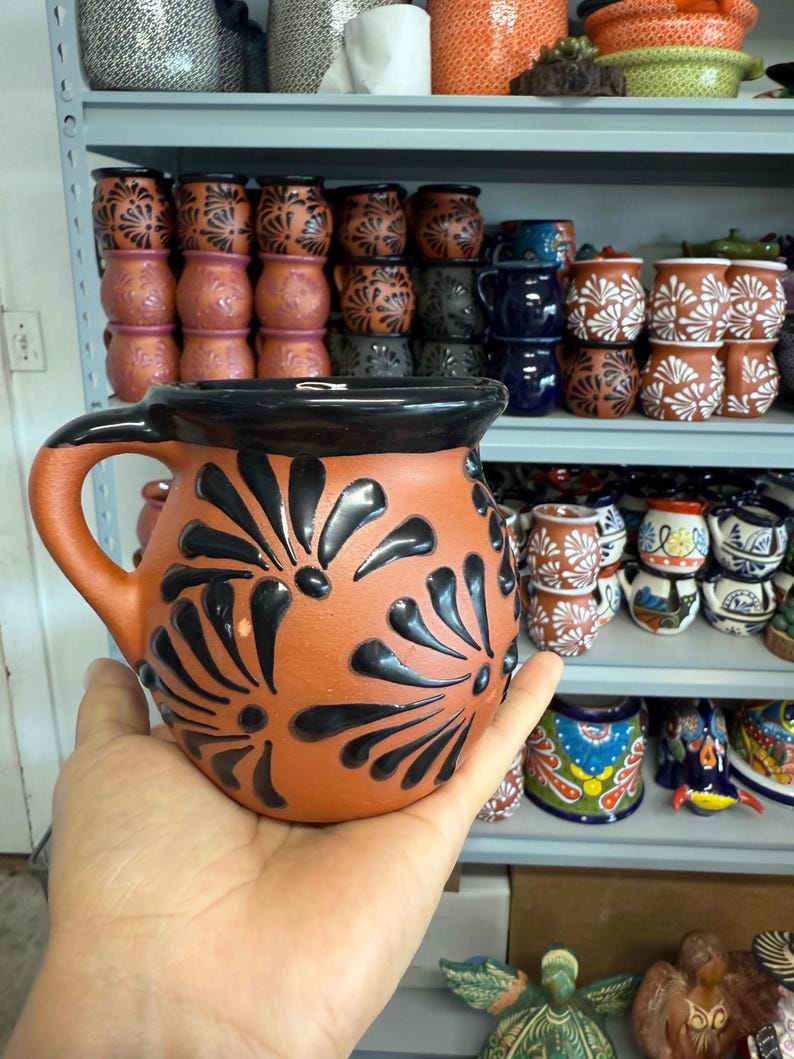 Puede incluir: Una jarra de terracota con un borde negro y dise&ntilde;os florales negros. La jarra tiene un asa y est&aacute; hecha de arcilla.