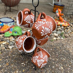 Puede incluir: Conjunto de seis tazas de terracota con dise&ntilde;os florales blancos, colgadas de un estante de metal negro. Las tazas est&aacute;n dispuestas en c&iacute;rculo, con las asas hacia afuera.