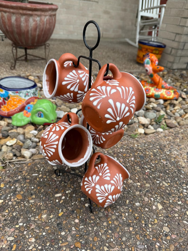 Puede incluir: Conjunto de seis tazas de terracota con dise&ntilde;os florales blancos, colgadas de un estante de metal negro. Las tazas est&aacute;n dispuestas en c&iacute;rculo, con las asas hacia afuera.