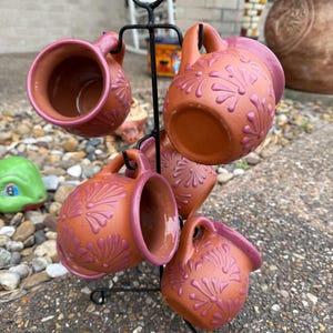 Puede incluir: Juego de seis tazas de terracota con dise&ntilde;os florales rosas, colgadas de un soporte de metal negro.