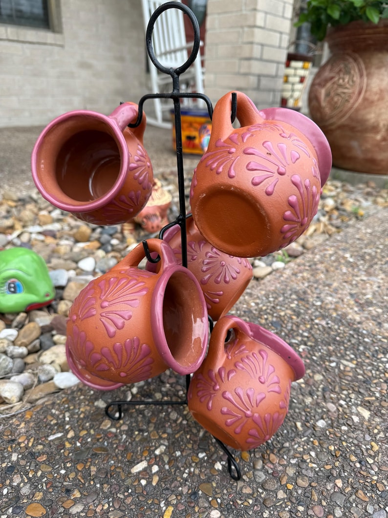 Puede incluir: Juego de seis tazas de terracota con dise&ntilde;os florales rosas, colgadas de un soporte de metal negro.