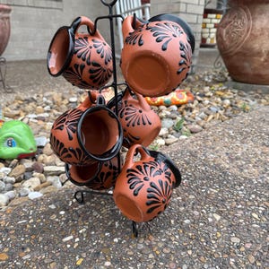 Puede incluir: Conjunto de seis tazas de terracota con dise&ntilde;os florales negros, expuestas en un soporte de metal negro. Las tazas tienen un aspecto r&uacute;stico y hecho a mano.