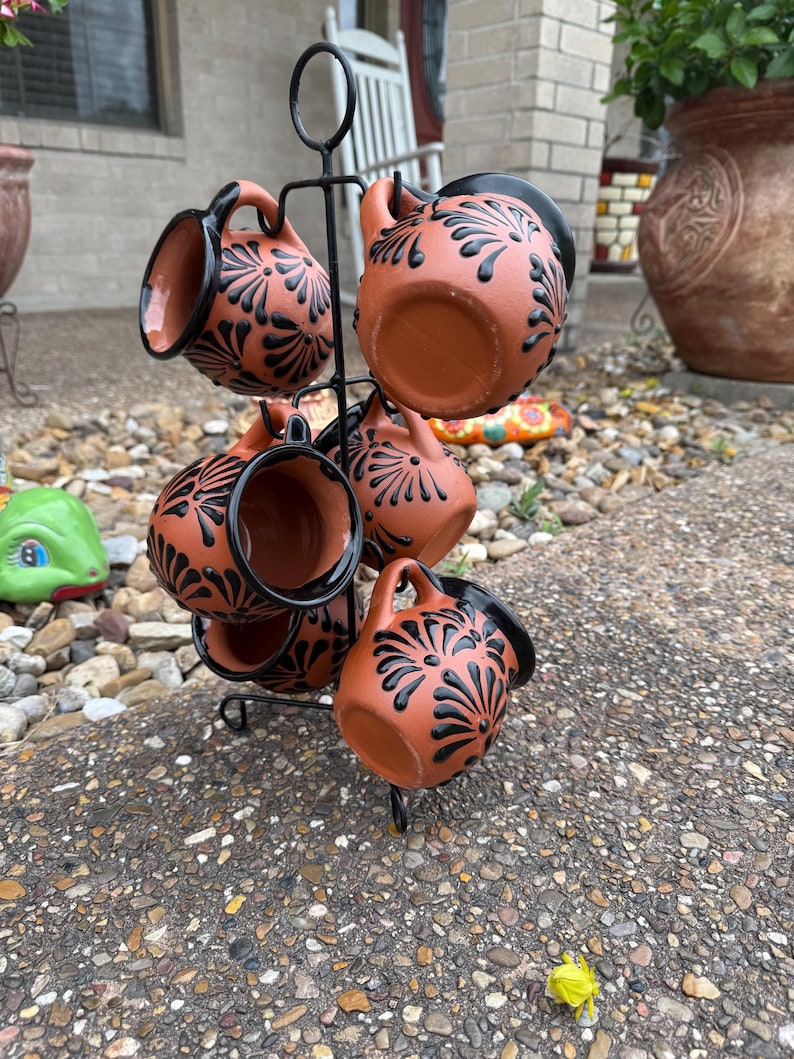 Puede incluir: Conjunto de seis tazas de terracota con dise&ntilde;os florales negros, expuestas en un soporte de metal negro. Las tazas tienen un aspecto r&uacute;stico y hecho a mano.