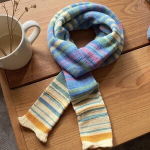 May include: A multi-coloured striped scarf in blue, pink, green, and yellow. The scarf is made of a soft, knitted material and is displayed on a wooden surface. The scarf is a cosy accessory.