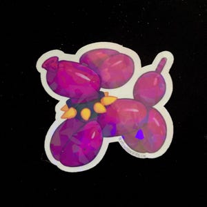 Op de afbeelding: Een holografische sticker van een roze ballondier in de vorm van een hond met een zwarte, met studs bezette halsband. De sticker heeft een glanzende, iriserende afwerking.