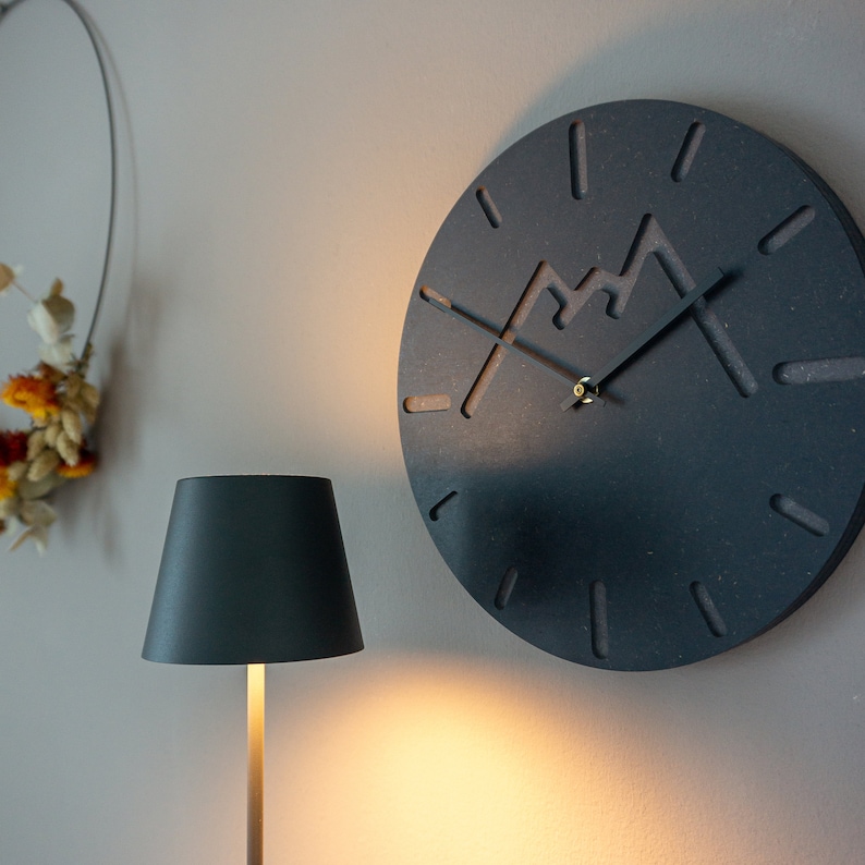 May include: A black clock with a mountain design and gold hands on a white wall. A black lamp with a white lampshade is in the foreground.