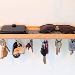 Op de afbeelding: Houten sleutelhouder met een plank. Een zwarte portemonnee en zonnebril liggen op de plank. Verschillende sleutels en sleutelhangers hangen eronder. De plank is ongeveer 30 cm lang.