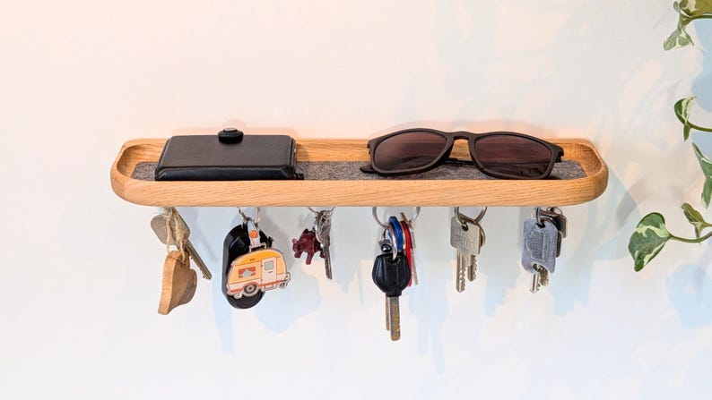 Op de afbeelding: Houten sleutelhouder met een plank. Een zwarte portemonnee en zonnebril liggen op de plank. Verschillende sleutels en sleutelhangers hangen eronder. De plank is ongeveer 30 cm lang.