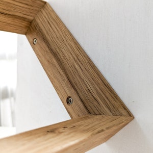 K&ouml;nnte beinhalten: Nahaufnahme einer Holzregal mit zwei Schrauben, die sie an einer wei&szlig;en Wand befestigen. Das Regal ist aus Naturholz mit einem Knoten und einer Maserung.