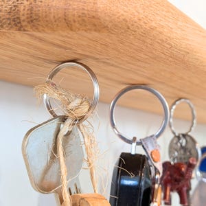 Op de afbeelding: Close-up van sleutels die aan een houten plank hangen. De sleutels zijn bevestigd aan metalen sleutelringen. E&eacute;n sleutelring heeft een natuurlijk touwdetail. Andere sleutelhangers zijn een zwarte fob, een rode olifant en een zilveren bedel. De plank is gemaakt van lichtgekleurd hout.