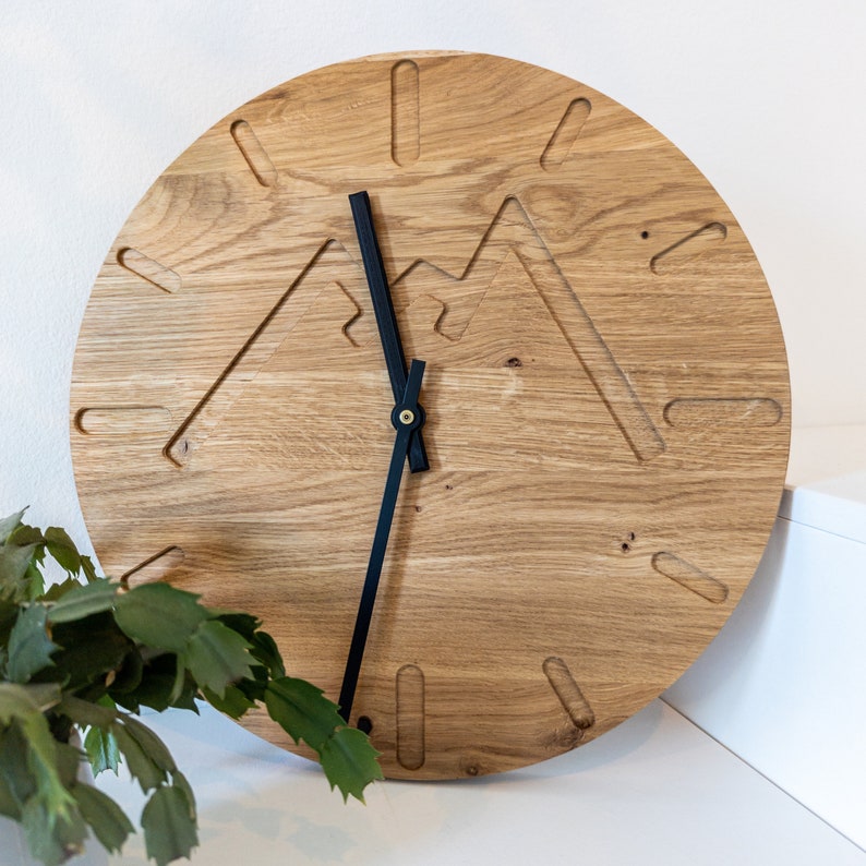 May include: A wooden wall clock with black hands and a mountain design. The clock face is made of light brown wood with a natural grain pattern.