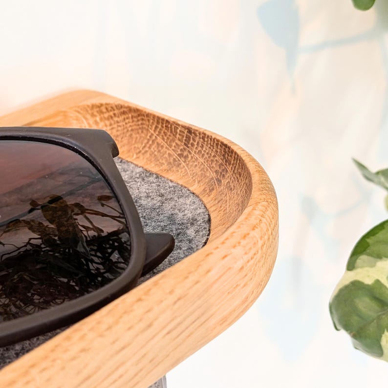 Op de afbeelding: Een houten zonnebrilhouder met een grijze vilt binnenkant. De houder heeft een gebogen ontwerp en is gemaakt van lichtgekleurd hout. Een zwarte zonnebril rust binnenin.